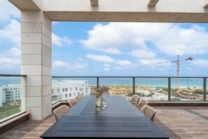 Un balcone con un tavolo apparecchiato per due persone, affaccia su una vista costiera con edifici e una gru all'orizzonte.
