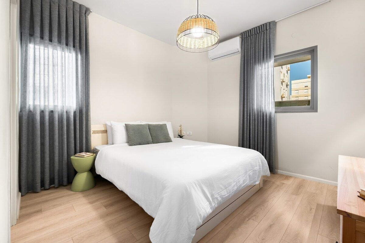 Ein Schlafzimmer mit einem Bett, einem Nachttisch, einer Lampe und einem Fenster mit Vorhängen.