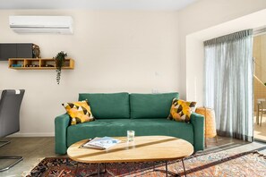 Un salon avec un canapé vert, une table basse, un téléviseur fixé au mur et une fenêtre avec des rideaux.