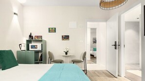 Un dormitorio con cama, escritorio, silla, una mesita con una planta y una puerta que da a otra habitación.
