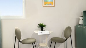 Une pièce avec une fenêtre, un tableau encadré, une petite table avec une plante et deux chaises.