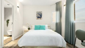 Une chambre à coucher moderne avec un grand lit, une table de chevet, une lampe fixée au mur et une fenêtre avec des rideaux.