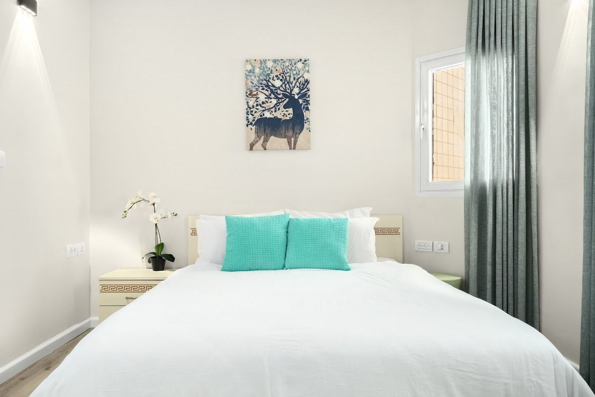 A bedroom with a bed, a nightstand, a potted plant, a window, and a wall-mounted artwork.