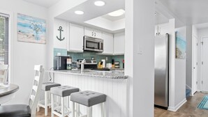 A modern kitchen with white cabinetry, a stainless steel refrigerator, and a bar area with stools. - Coastal Penthouse Paradise Indoor Heated Pool Marsh View (Myrtle Beach)