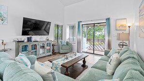 A living room with a large flat-screen TV, a green sofa, a wooden coffee table, and a sliding glass door leading to an outdoor area. - Coastal Penthouse Paradise Indoor Heated Pool Marsh View (Myrtle Beach)
