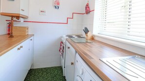 Une cuisine compacte avec des meubles blancs, un plan de travail en bois, un lave-linge et une fenêtre avec des stores.