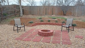 A backyard with a circular fire pit surrounded by red bricks, two chairs, and a wooden fence in the background. - Spacious 6-bedroom house with WiFi, fitness room in peaceful Thornton (Thornton)