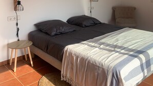 Une chambre avec un lit, un petit tapis rond en fibres tressées, un petit tabouret en bois et une lampe fixée au mur.