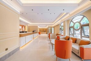 Un hall de hotel con una larga recepción, zona de estar con sillas anaranjadas y ventanas amplias.
