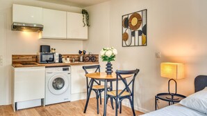 A compact kitchen with a washing machine, microwave, and wooden countertop. There is a dining table with chairs and a vase of flowers. A lamp is on a bedside table next to a bed.