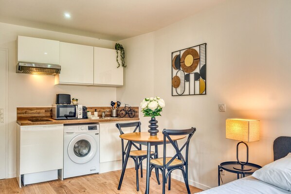 A compact kitchen with a washing machine, microwave, and wooden countertop. There is a dining table with chairs and a vase of flowers. A lamp is on a bedside table next to a bed.