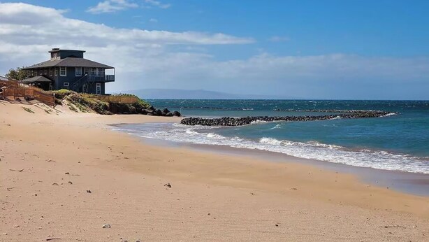 Une plage avec un bâtiment sur la gauche, une digue rocheuse dans l’eau et un ciel dégagé.