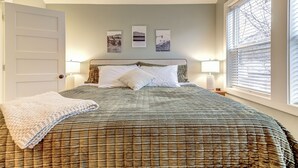 Un dormitorio con cama, almohadas, una colcha, decoración en la pared y una ventana con persianas.