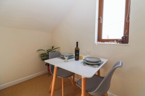 A small dining area with a white table, two grey chairs, a bottle of wine, and a window with curtains. - Priory Barn Apartment (Street)