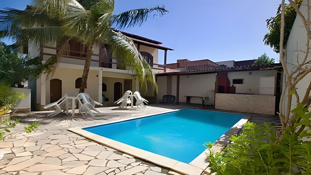 A swimming pool with stone tiles, surrounded by palm trees and a building with a tiled roof. - Porto D Aldeia - Cabo Frio RJ - 1 (Sao Pedro da Aldeia)
