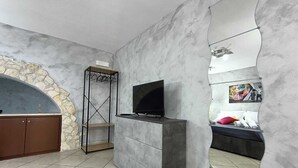 Un salon moderne avec un mur en arc de pierre, un téléviseur à écran plat posé sur un meuble, un portant à vêtements et un lit agrémenté d’une affiche colorée.