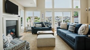 A spacious living room with a fireplace, comfortable seating, large windows, and a flat-screen TV.