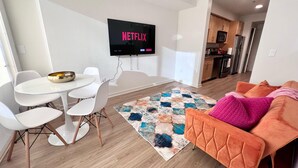 Un salon moderne avec une table à manger blanche, des chaises blanches, un canapé orange et un tapis coloré. Un téléviseur à écran plat fixé au mur affiche le logo de Netflix.