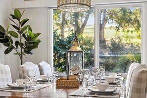 Une table à manger dressée avec de la verrerie et de l’assiette, une lanterne décorative, et une vue sur un jardin par de grandes fenêtres.
