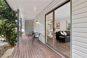 A covered patio with a wooden deck, a sliding glass door leading to a living area with a sofa and a small round table. - Holliday Haven (Lorne)
