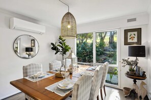 A dining area with a wooden table set for four, a hanging light fixture, and a view of a garden through large windows. - Fair Waves (Lorne)