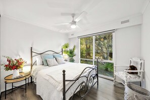 Een slaapkamer met een bed, een nachtkastje met een lamp, een stoel en uitzicht op groen door een groot raam.