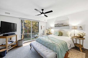Een slaapkamer met een bed, een televisie, een plafondventilator en een raam met uitzicht op bomen.