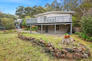 A two-story house with a covered porch, a stone wall, and a tree stump with potted plants. - Holliday Haven (Lorne)