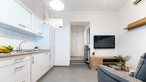 Une cuisine moderne avec des armoires blanches, un comptoir en granit et un saladier rempli de fruits. On y trouve une table à manger en bois avec des chaises et un canapé gris. Un téléviseur à écran plat est fixé au mur.