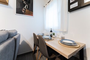 Un comedor con una mesa de madera dispuesta para cuatro personas, incluyendo platos, vasos y una botella de vino. Se observa un sofá gris al fondo y una ventana con cortinas ligeras.