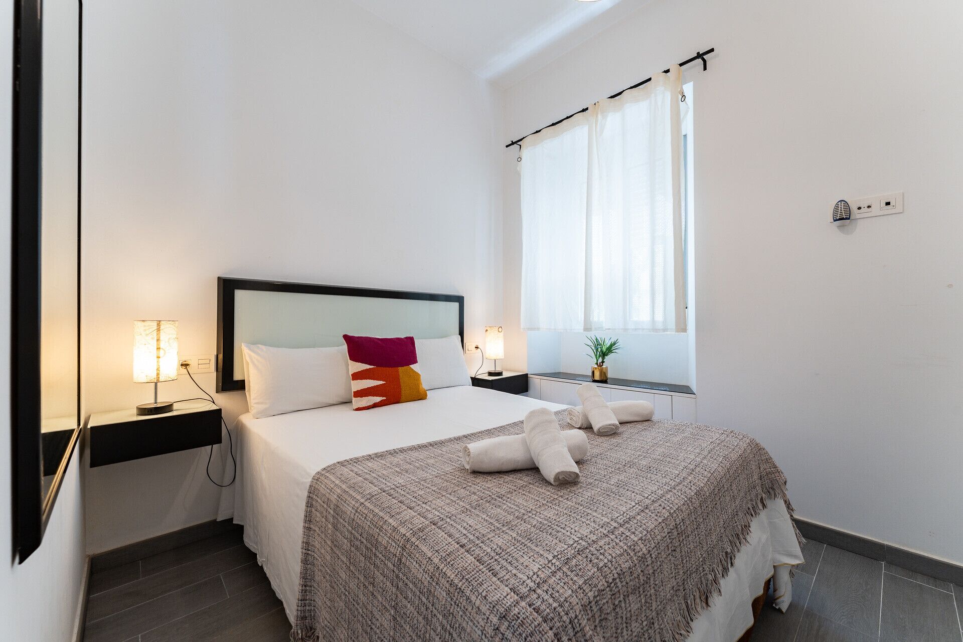 Une chambre à coucher bien rangée, avec un lit, des lampes de chevet, une fenêtre avec des rideaux et une petite plante sur le rebord de la fenêtre.
