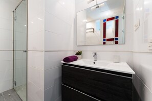 Un baño moderno con cabina de ducha de vidrio, un lavabo blanco sobre un mueble de tocador oscuro y un espejo con marco decorativo rojo y azul.