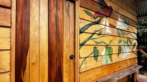 A wooden building with a door labeled "Joe," a mural of a hummingbird and flowers, and a wooden bench outside. - Pura Chata Ecolodge (San Juan Bautista)