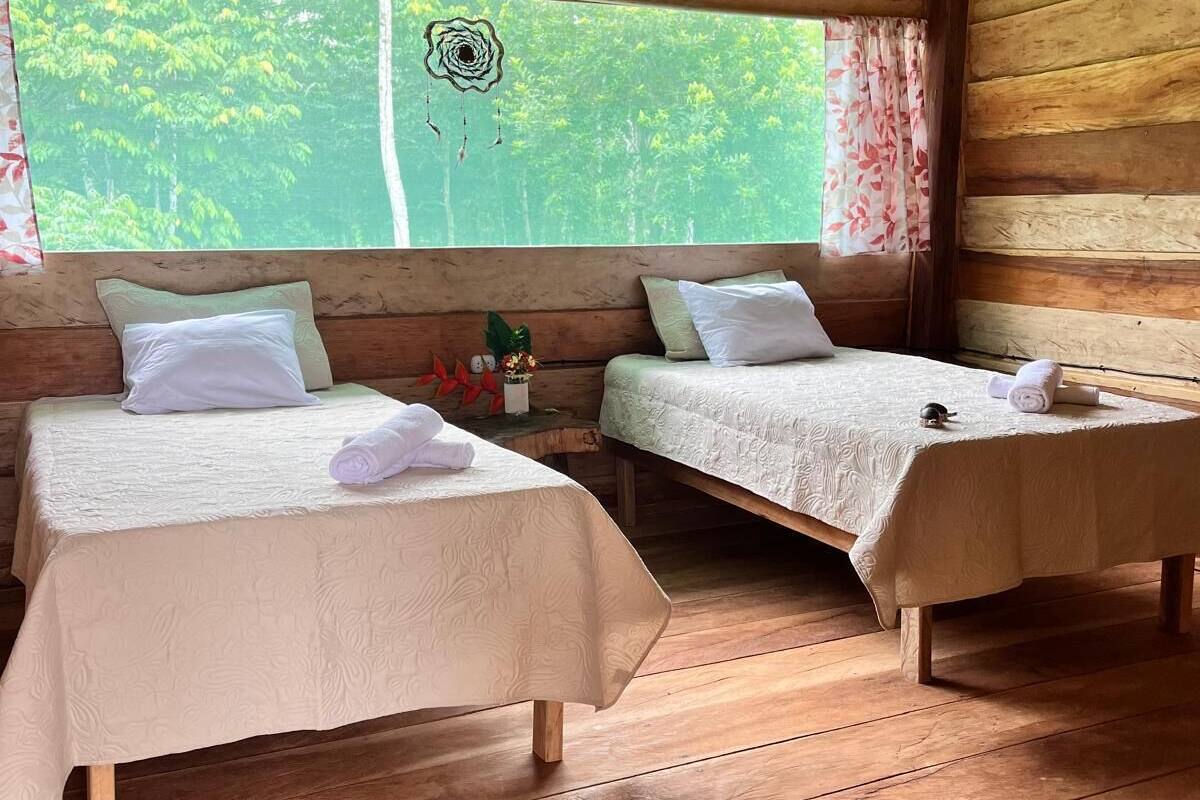 A cabin with two wooden beds, a window with a view of trees, and a dreamcatcher hanging on the wall.