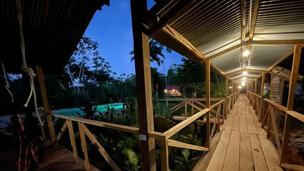 A wooden deck with railings leading to a pool area, surrounded by lush greenery. - Pura Chata Ecolodge (San Juan Bautista)