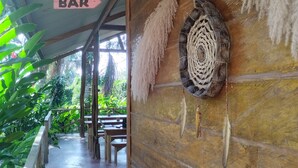 A wooden corridor with a sign indicating a bar, a woven wall hanging, and a concrete pathway leading to outdoor seating. - Pura Chata Ecolodge (San Juan Bautista)