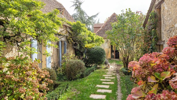 A narrow stone pathway flanked by lush greenery and traditional buildings with tiled roofs.
