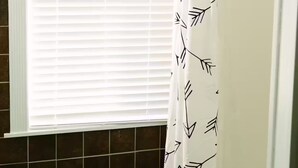 Une salle de bain avec une fenêtre, des stores blancs et un rideau de douche orné d’un motif de feuilles.