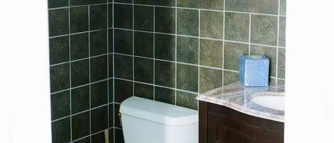 A bathroom with a toilet, dark tiled walls, a wooden shelf, and a vanity with a mirror.