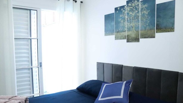 Une chambre à coucher comprenant un lit, une tête de lit, une fenêtre avec des stores et trois tableaux accrochés au mur.