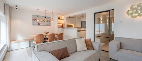 A modern living room with a sofa, a dining area with chairs, and a kitchen in the background.