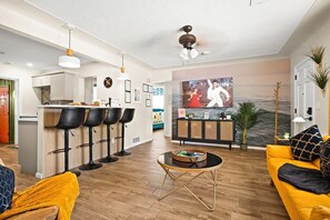 Una sala moderna con un sofá amarillo, una zona de comedor con taburetes de bar, un televisor montado en la pared y un ventilador de techo.