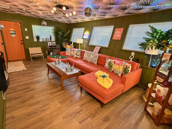 A living room with a sectional sofa, a coffee table, a wooden floor, a ceiling with a disco ball, and a door.