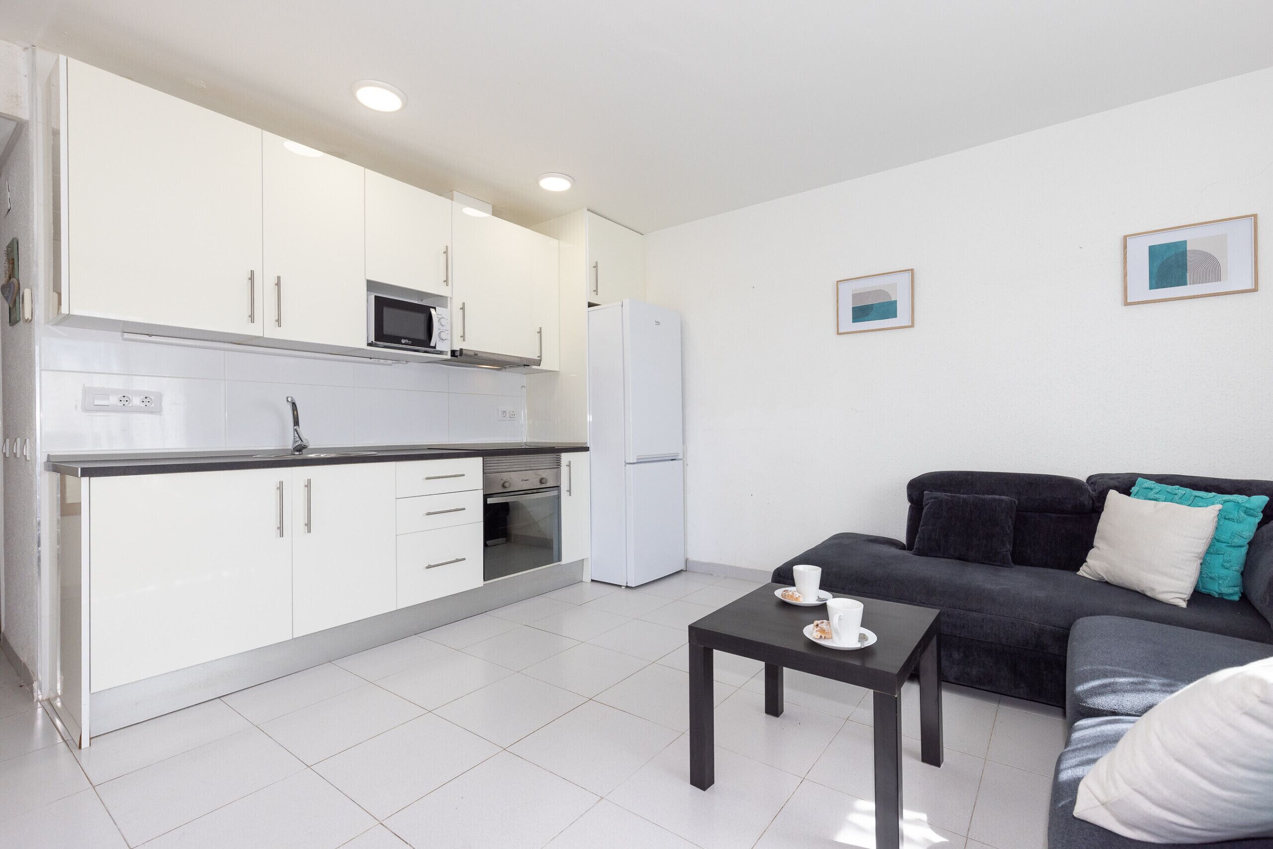Una cocina moderna con armarios blancos, un microondas y un refrigerador. Hay un sofá de color oscuro con cojines y una pequeña mesa con tazas y platillos.