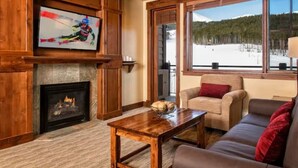A cozy living room with a fireplace, a television mounted on a wooden wall, a large window with a snowy view, and a comfortable sofa. - 4th of July in Breckenridge at Grand Lodge on Peak 7 (Breckenridge)