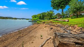 A lakeside beach with a wooden dock, a large tree, and a house with a stone facade. - Lakefront Oasis- Steps to Water- Private Boat Ramp (Dandridge)