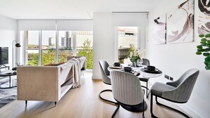 A modern living room with a sofa, dining table, and chairs. There are large windows with a view of buildings and greenery.