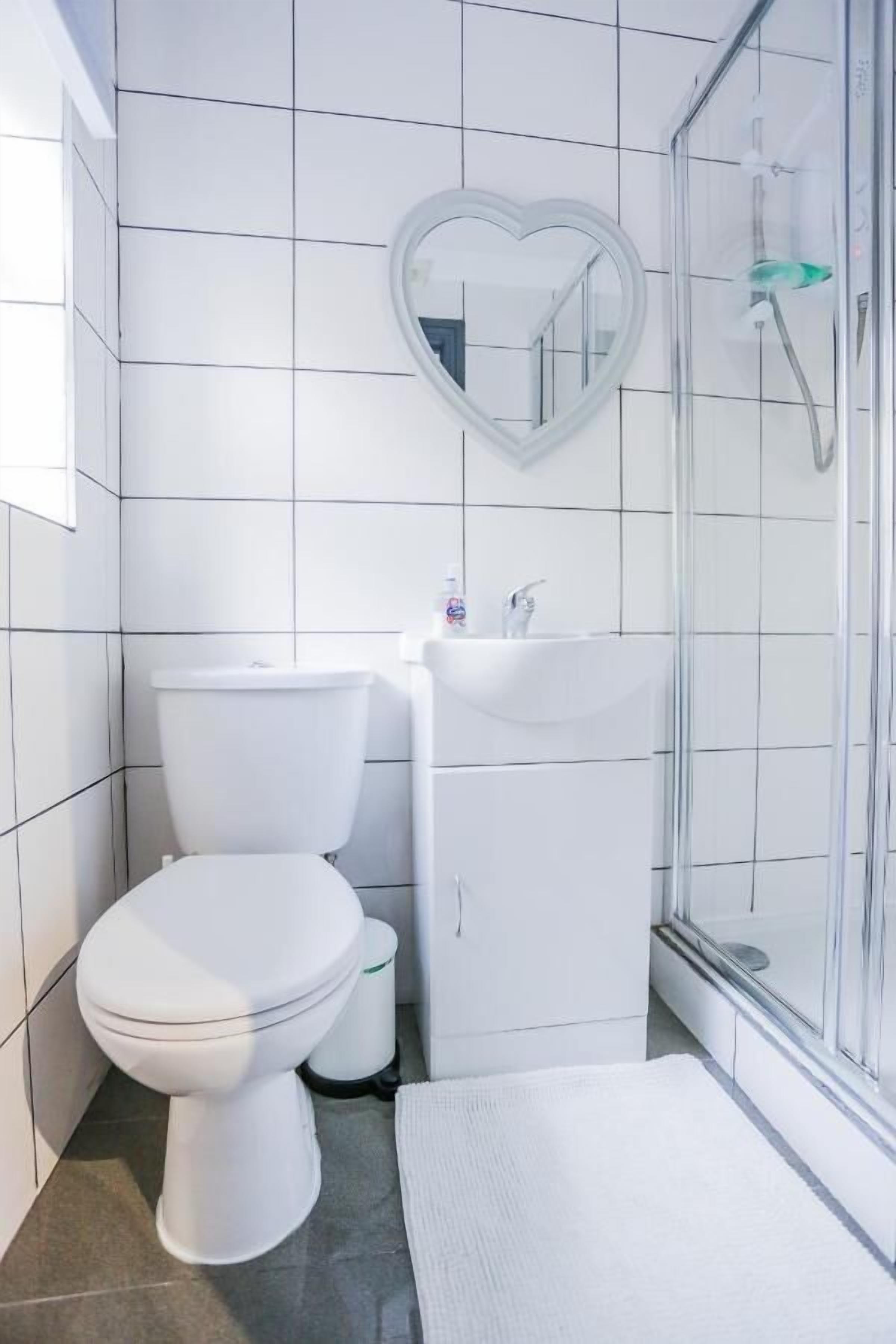 A modern bathroom with white tiles, a heart-shaped mirror, a toilet, a sink, and a shower enclosure.