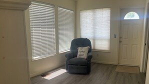 A room with a gray armchair, a window with blinds, a door, and a vent on the floor.