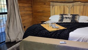 A wooden cabin room with a bed, a small table, and a window with curtains. - Hidden in East Texas Piney Woods - Rustic Western Cabin - Canton 1st Monday (Mineola)